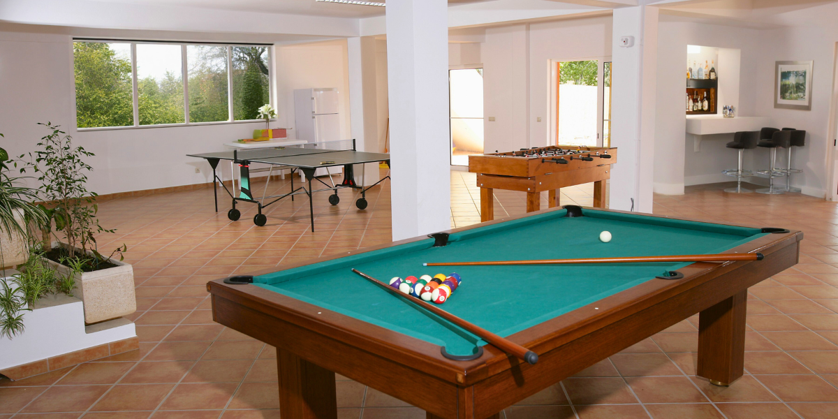 Well lit basement with windows with a pool table, ping pong table, foosball table, and a bar
